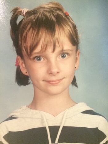 A seven or eight year old Rebecca in a striped shirt with long, straight bangs and her hair in some hard-to-describe pony tails that form a sort of headband.